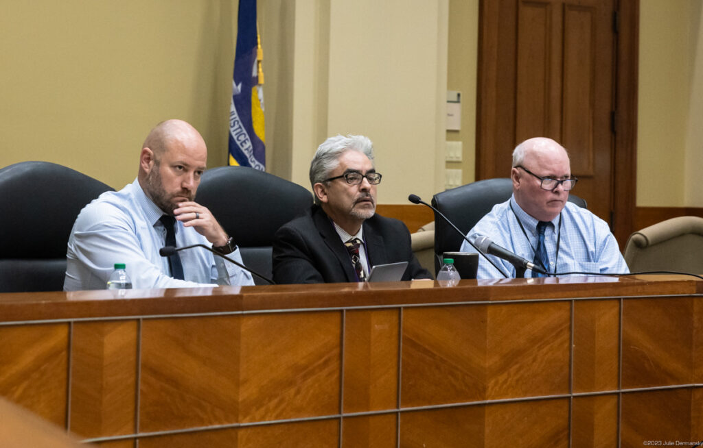 Three men in suits sit behind microphones at a wood-paneled dais.