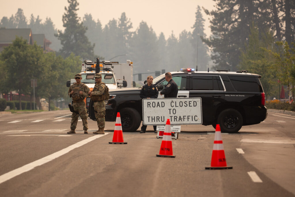 Two police officers and two military members stand in front of a black SUV with orange traffic cones and a 'road closed to through traffic' sign. Wildfire smoke hangs over evergreens behind them.