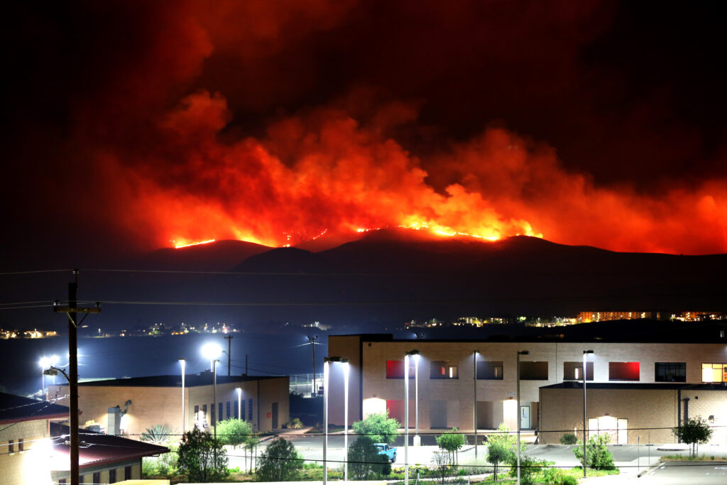 Red wildfire and smoke light up the hills at night beyond brightly lit beige government buildings and parking lots.