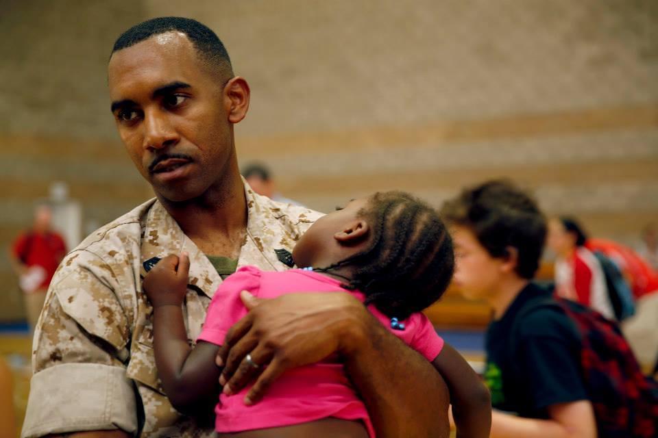 A father in beige military fatigues carries his sleeping daughter in an evacuation shelter.