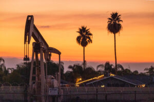 Oil pumpjack, a house with holiday lights, and palm trees set against an orange sky at sunset.