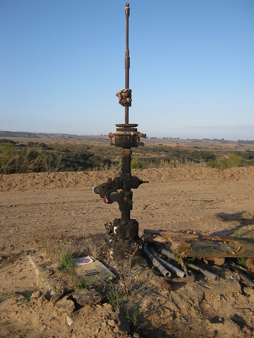 A tall rusty brown metal pipe with valves juts out of the dry soil, with a road, arid landscape, and blue sky beyond. 