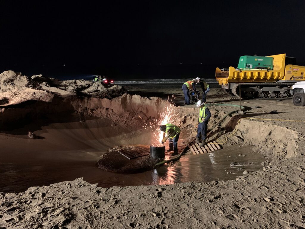 Workers in yellow safety vests weld, oversee, and run machinery to close an oil well, at night in a wide dirt hole.