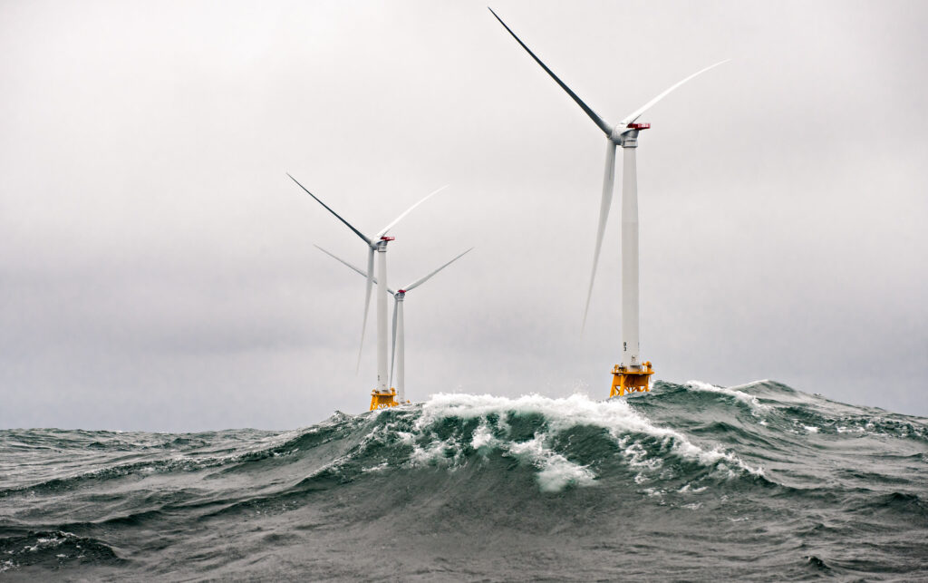 Heavy seas engulf three white wind turbines with yellow bases on a cloudy day.