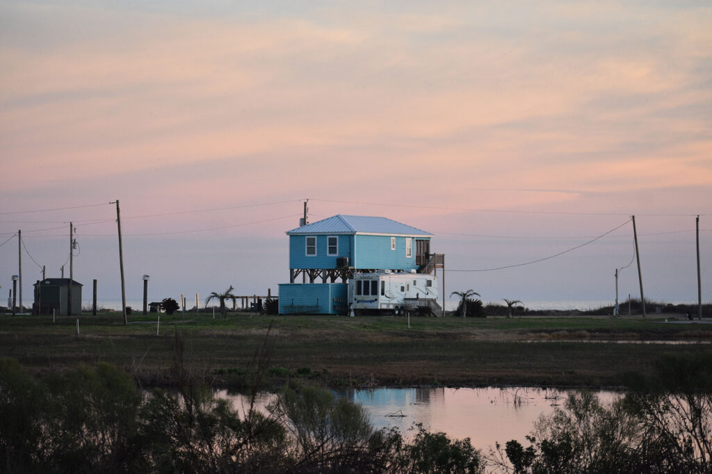 Bright turquoise house on stilts with water reflecting in the foreground at sunset.