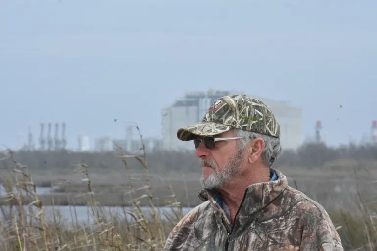 John Allaire, looking left and wearing camo baseball cap and jacket, photographed with fossil fuel infrastructure behind. 