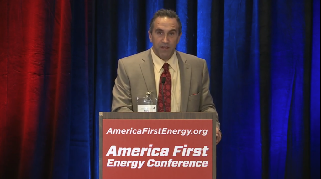 Marc Morano in a tan suit with red tie at a podium with red sign reading 'AmericafirstEnergy.org America First Energy Conference'