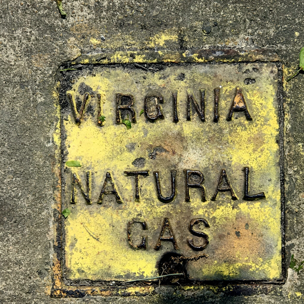 On cement, a metal utility cover painted yellow and reading 'VIRGINIA NATURAL GAS.'