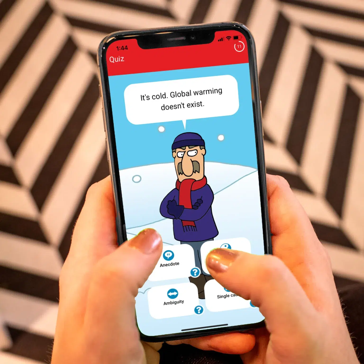 A person is holding a cell phone showing an image from the Cranky Uncle game. The cranky uncle is standing outside in the snow and saying, "It's cold. Global warming doesn't exist." Buttons underneath give the user options to identify the type of misinformation.