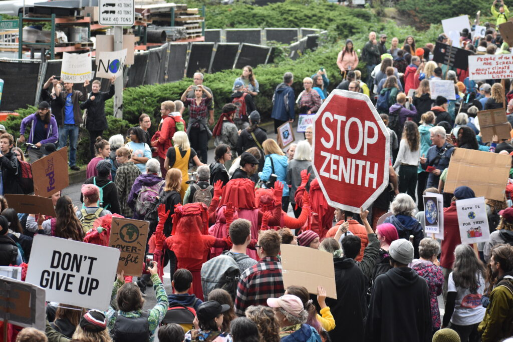 Dozens of people are visible in the photo, some holding signs. The messages on the signs include "Stop Zenith" and "Don't Give up."