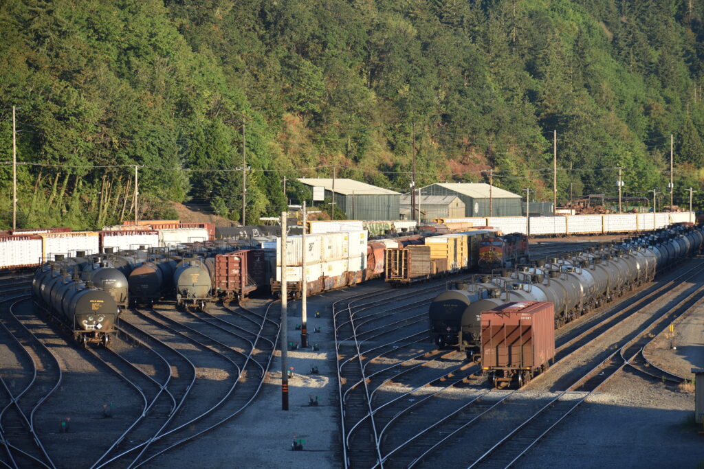 Oil trains on tracks point toward the camera. Behind them are buildings and trees.