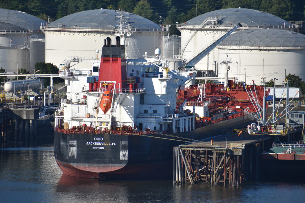 The tanker says "Ohio" and "Jacksonville, FL." It is near a pier and a lot of equipment, with the storage tanks just beyond.