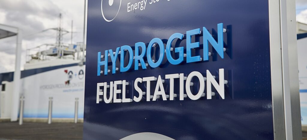 A sign for a hydrogen fuel station is in the foreground in front of infrastructure