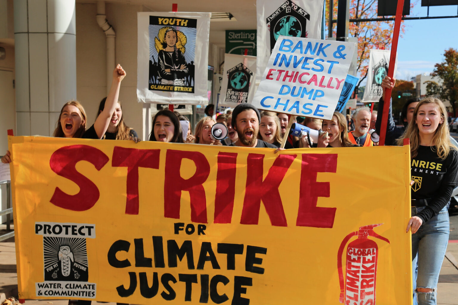People holding signs, including one that says "Bank & Invest Ethically Dump Chase," stand behind a banner that says, "Strike for climate justice"