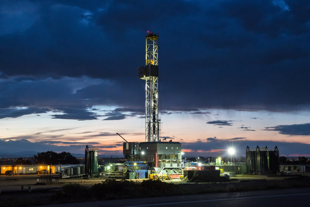 Dark clouds hang over the metal tower of a fracking drill rig lit up at twilight.