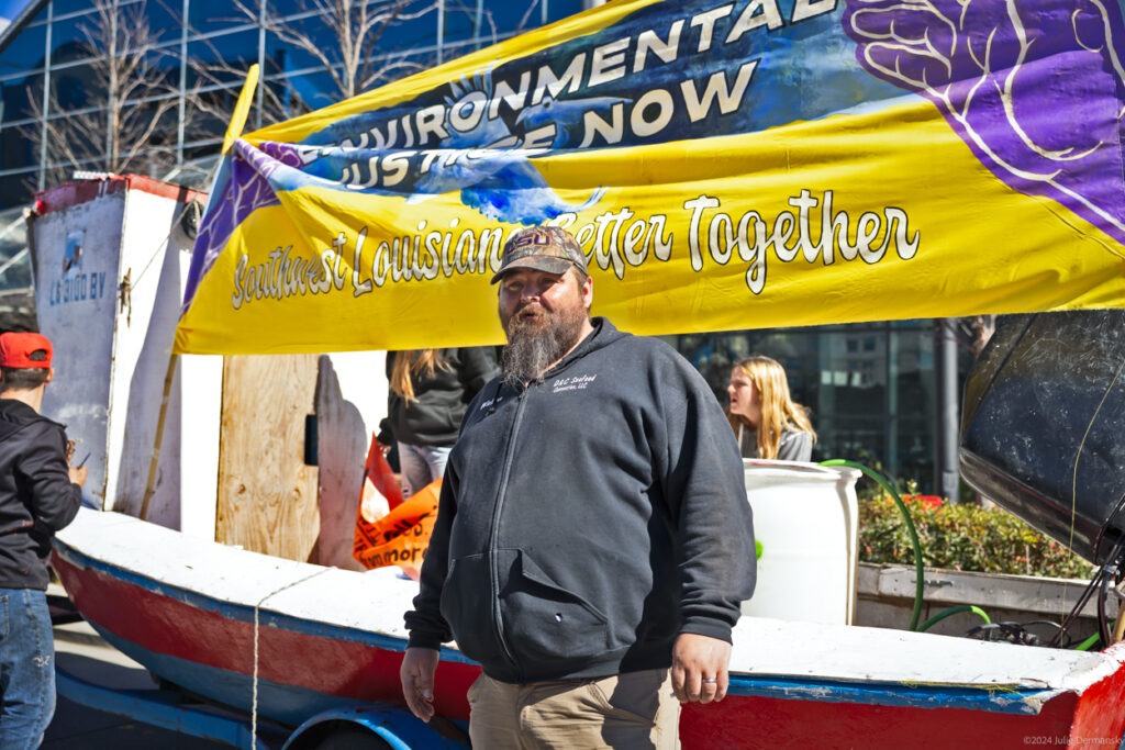Travis Dardar is wearing a gray hoodie that says "D&C Seafood" and an LSU baseball cap, standing beside a boat with a sign that says, "Environmental Justice Now: Southwest Louisiana Better Together"