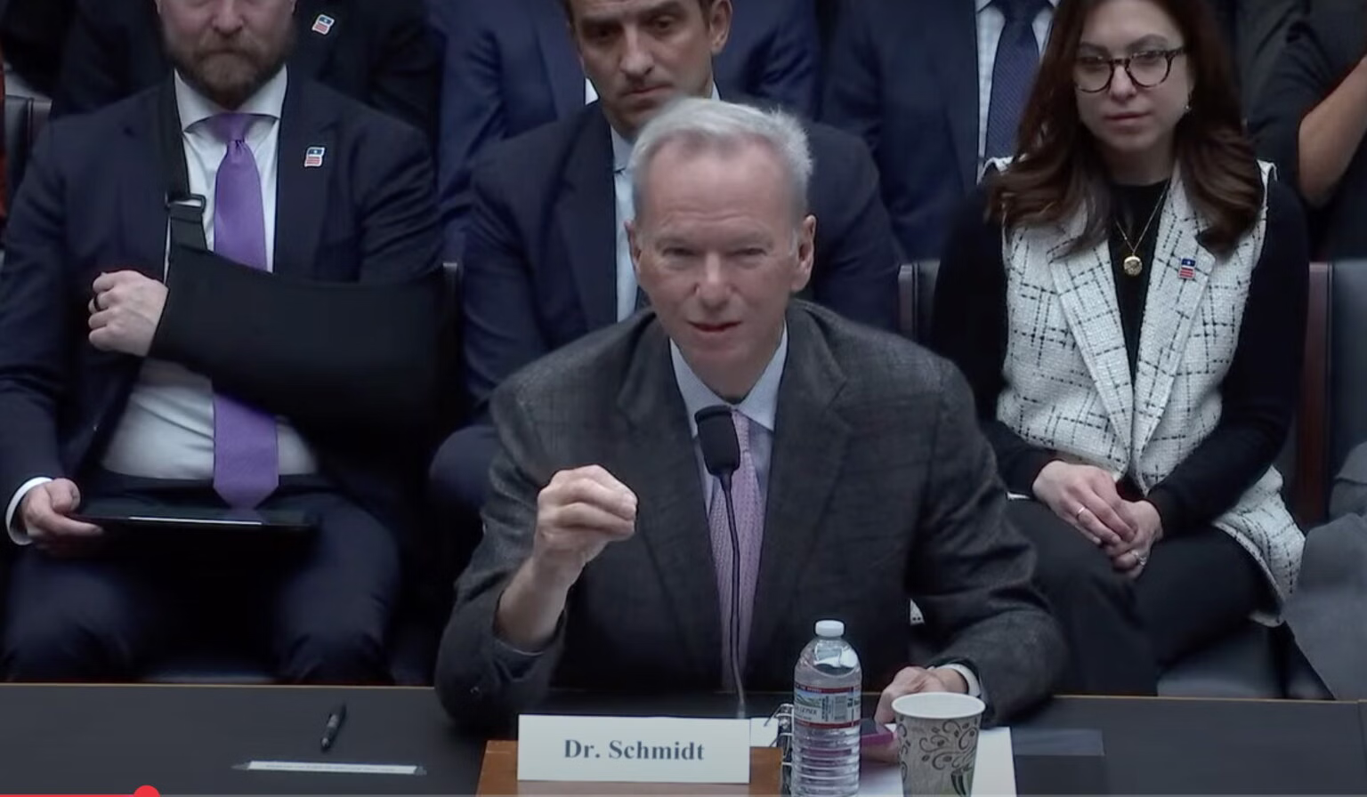 Former Google CEO Dr. Eric Schmidt testifying before Congress, sitting in a suit before a microphone with a water bottle. A row of men and women sit behind him.