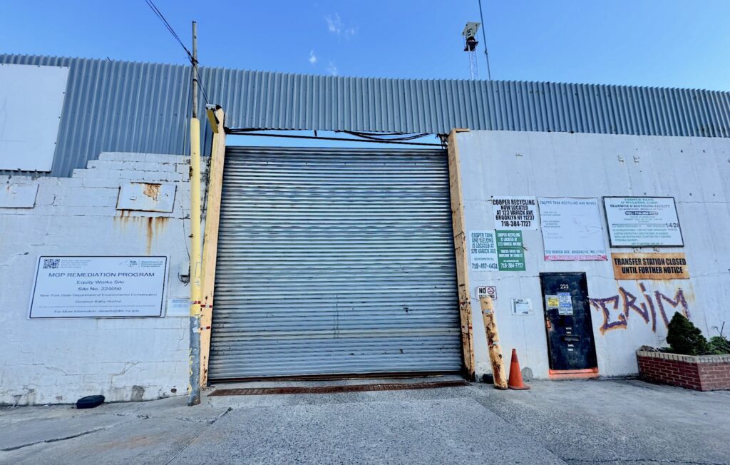 The gate to the Equity Works Gas Site in Brooklyn New York. Among several signed affixed to the dingy white brick walls on either side of the closed rolldown gate, one states it is in a state environmental remediation project.