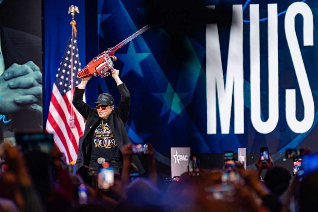 Elon Musk holding a chainsaw overhead onstage at the Conservative Political Action Conference (CPAC) 2025, with an American flag behind him and his last name in large white letters on a banner to his right.