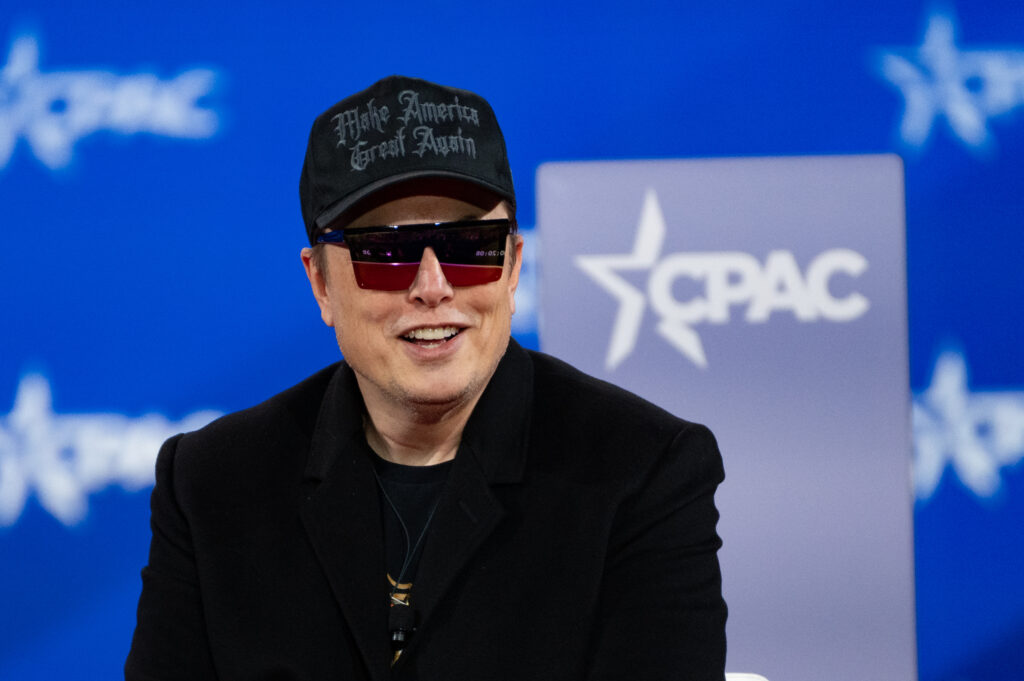 Elon Musk wearing a black 'Make America Great Again' baseball cap, seated, with a CPAC logo behind him on a gray podium and blue background.