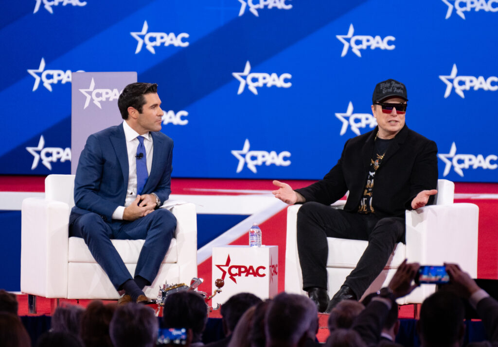 Elon Musk, right, sitting on the dais with a white male moderator in a suit, left, on stage at CPAC. CPAC logos in white on blue background behind them.