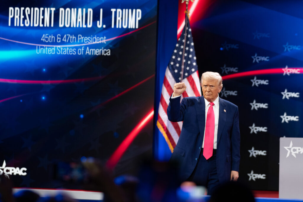 President Donald Trump raising a fist in front of an American flag on stage right, with 'President Donald J Trump, 45th and 47th President of the United States of America' projected in white on blue and black background.