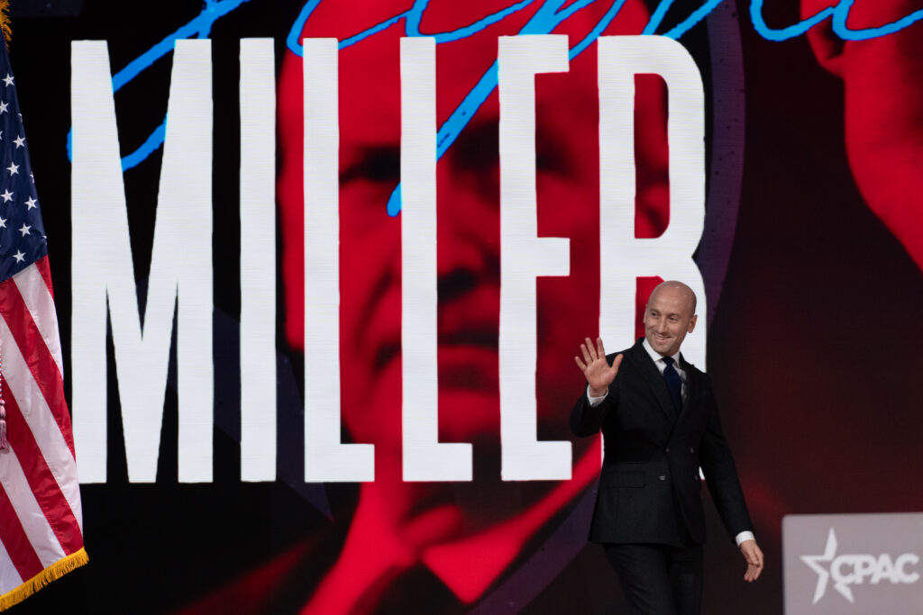 Stephen Miller walking right across a stage toward a podium labeled CPAC, with 'MILLER' in large white letters projected behind him.