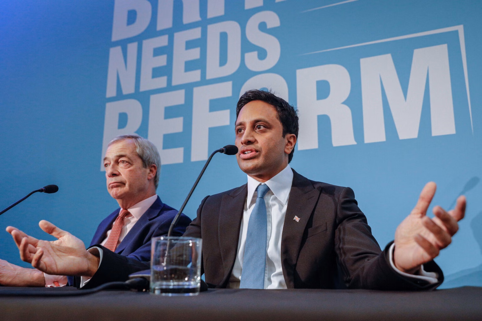 Reform UK leader Nigel Farage, left, and chair Zia Yusuf, right, sit at microphones and a black covered table with pale blue background and the words 'Britain needs reform.'