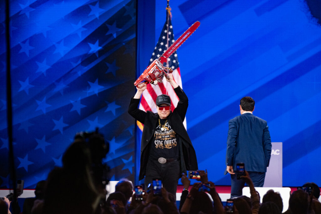 Elon Musk holding a chainsaw overhead onstage with an American flag behind him and a man in a suit facing the blue backdrop to his right.
