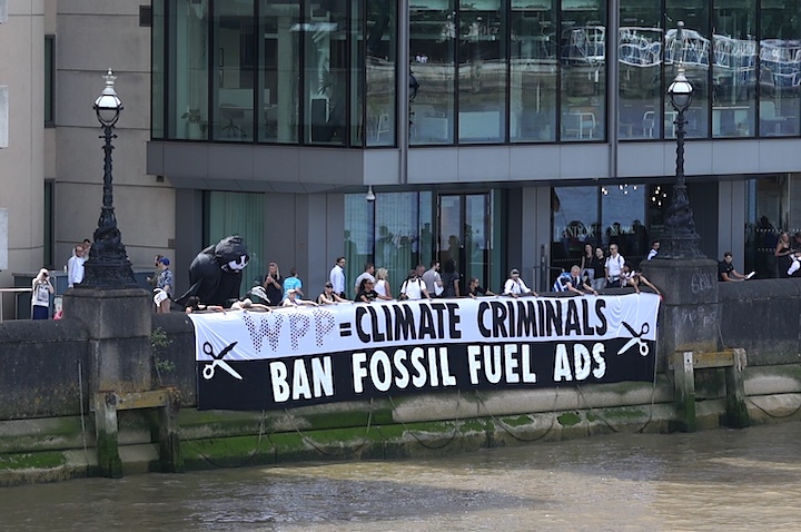 Several activists holding a fifteen foot long banner over the Thames Embankment, in front of the headquarters of WPP. The banner reads WPP are climate criminals, ban fossil fuel advertising
