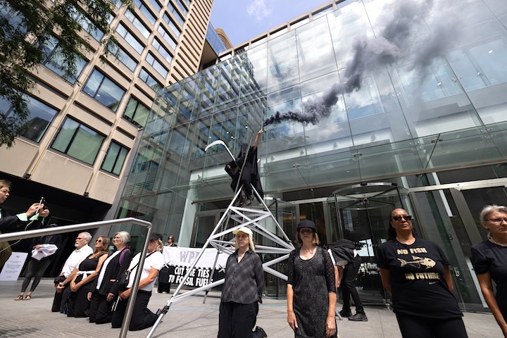 Several activists protesting outside London offices of ad agency WPP. They are dressed simply in black and white, and standing in an arc in front of a banner that says wpp cut the ties to fossil fuels. Behind them, someone dressed up as the grim reaper stands atop a scaffolding that resembles an oil rig, holding a cylinder that is emitting a plume of black smoke.