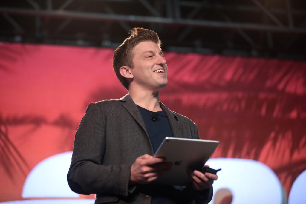 Alex Epstein smiles in a suit jacket holding an iPad and looking right, with a red background.