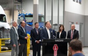 Lee Zeldin, center, stands at a podium and Chris Wright, left, are joined by other government officials and staff in a truck manufacturing facility.