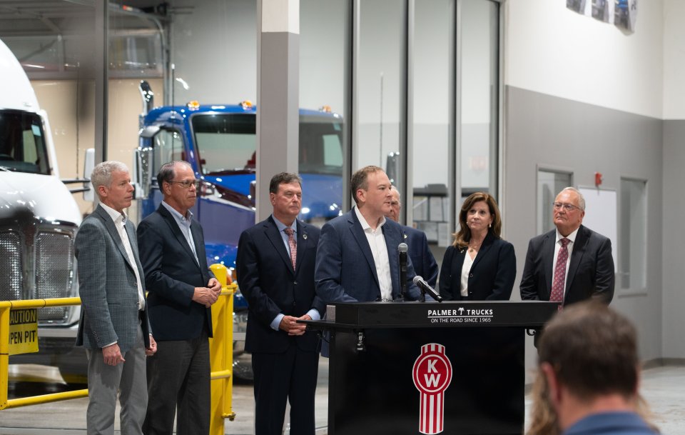 Lee Zeldin, center, stands at a podium and Chris Wright, left, are joined by other government officials and staff in a truck manufacturing facility.