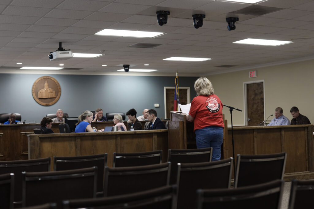 A blond woman in a red t-shirt reading 'Citizens for Rural Coweta' testifies at a podium in a low-ceilinged room before a panel of people representing the Coweta County Commission and administrators in seats and behind wood paneling look down.