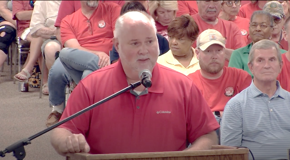 Man in red shirt speaking into a microphone while standing at a podium. Behind him the seats are filled with people.