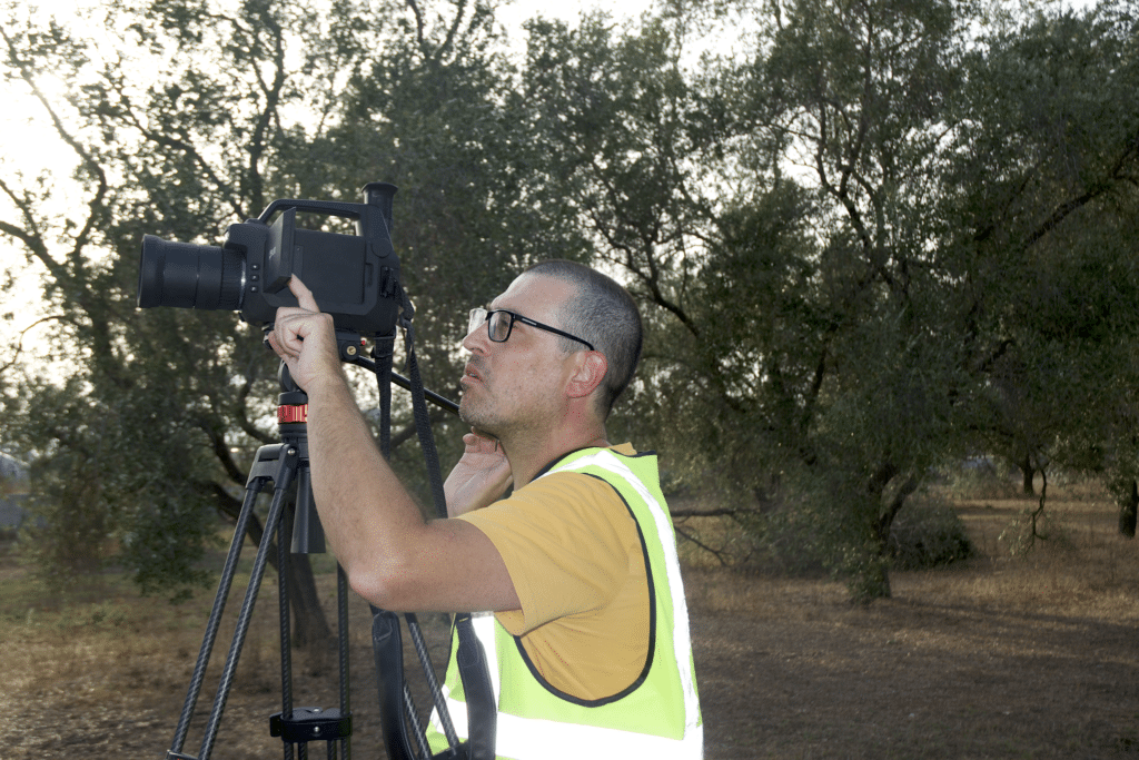 Man in yellow safety vest aiming a large camera on a tripod towards something beyond the frame of the photo.