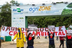 During the COP30 climate summit in Brazil, people are holding signs and a banner protesting environmental pollution by agriculture. They are standing in front of the Agrizone, a special event venue sponsored by agribusiness and located near the main conference venue.