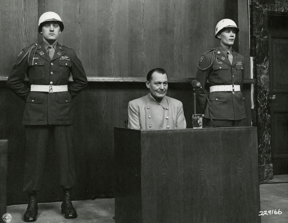 A man sits in a witness booth with guards on either side.