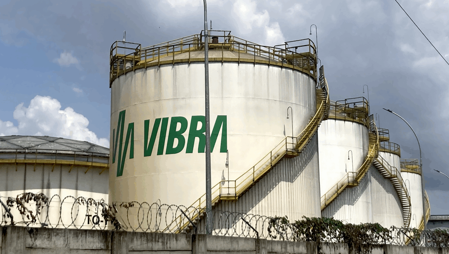 A line of giant white fuel silos, one behind the other receding to the right. The Vibra logo painted in green is visible on the front silo.
