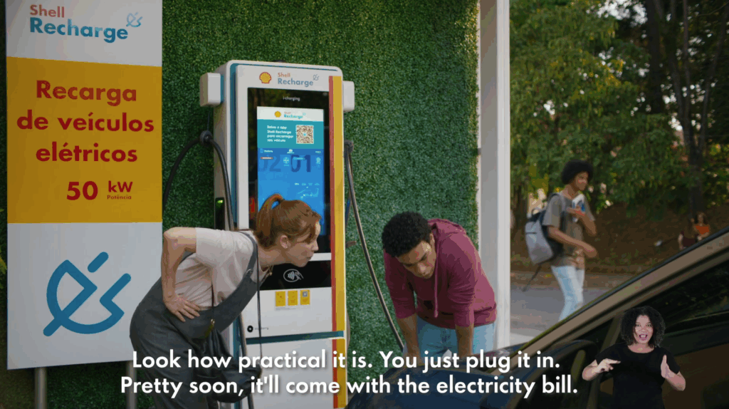 A video still of a Shell ad in Brazil. A man and a woman at a Shell station charging point are about to attach the electric vehicle charging cable to their car. An English subtitle at the bottom of the screen, Look how practical it is. You just plug it in. Pretty soon, it'll come with the electricity bill.