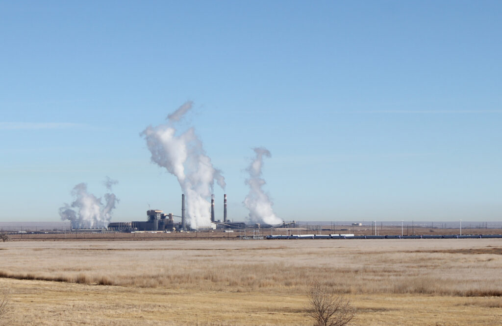 The coal-fired power plant, the Comanche Generating Station, with its smokestacks emitting white smoke in the distance on a flat beige plain and a blue sky.