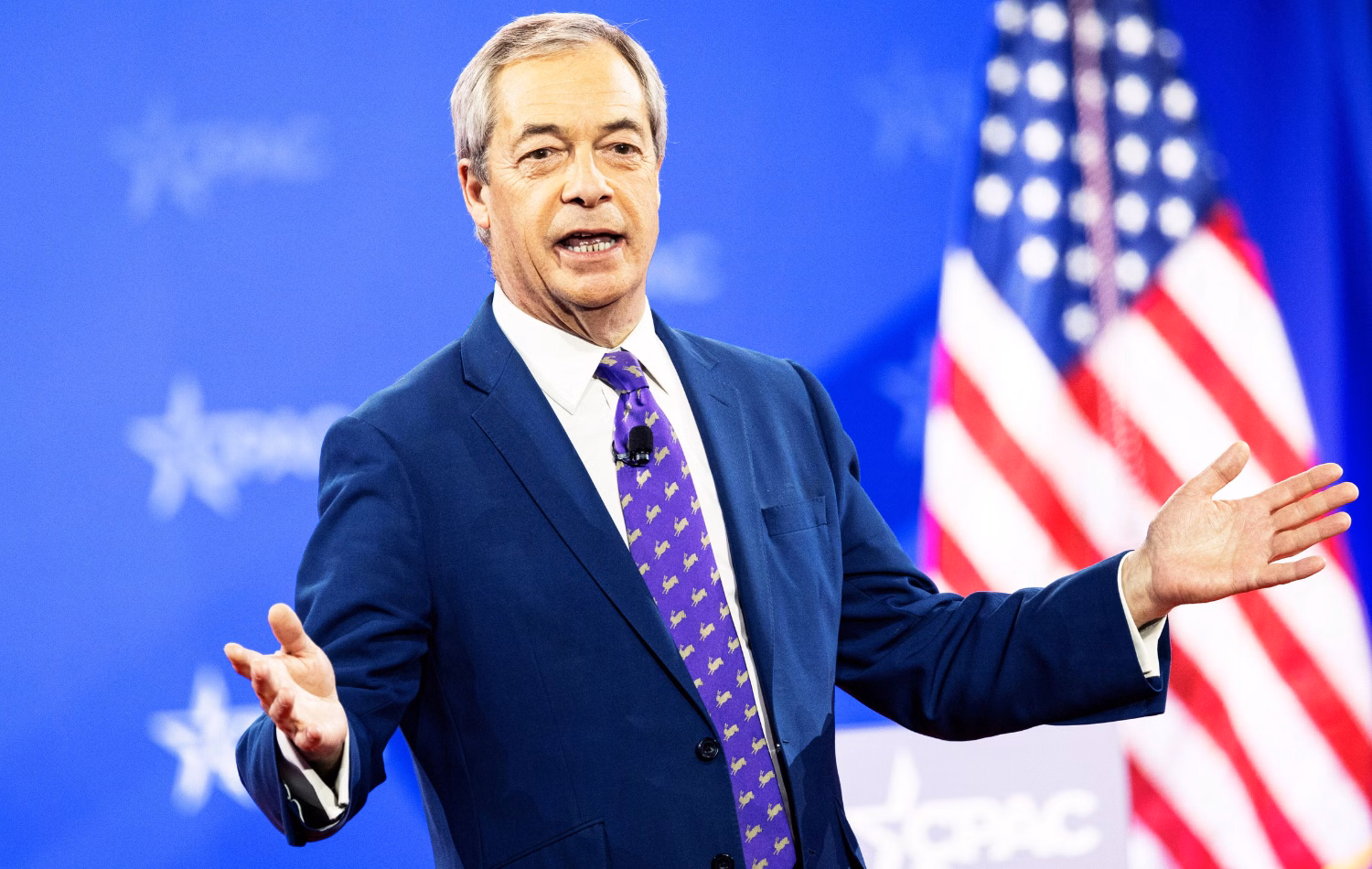 Reform UK leader Nigel Farage speaking at CPAC against a blue background and American flag in February 2025.
