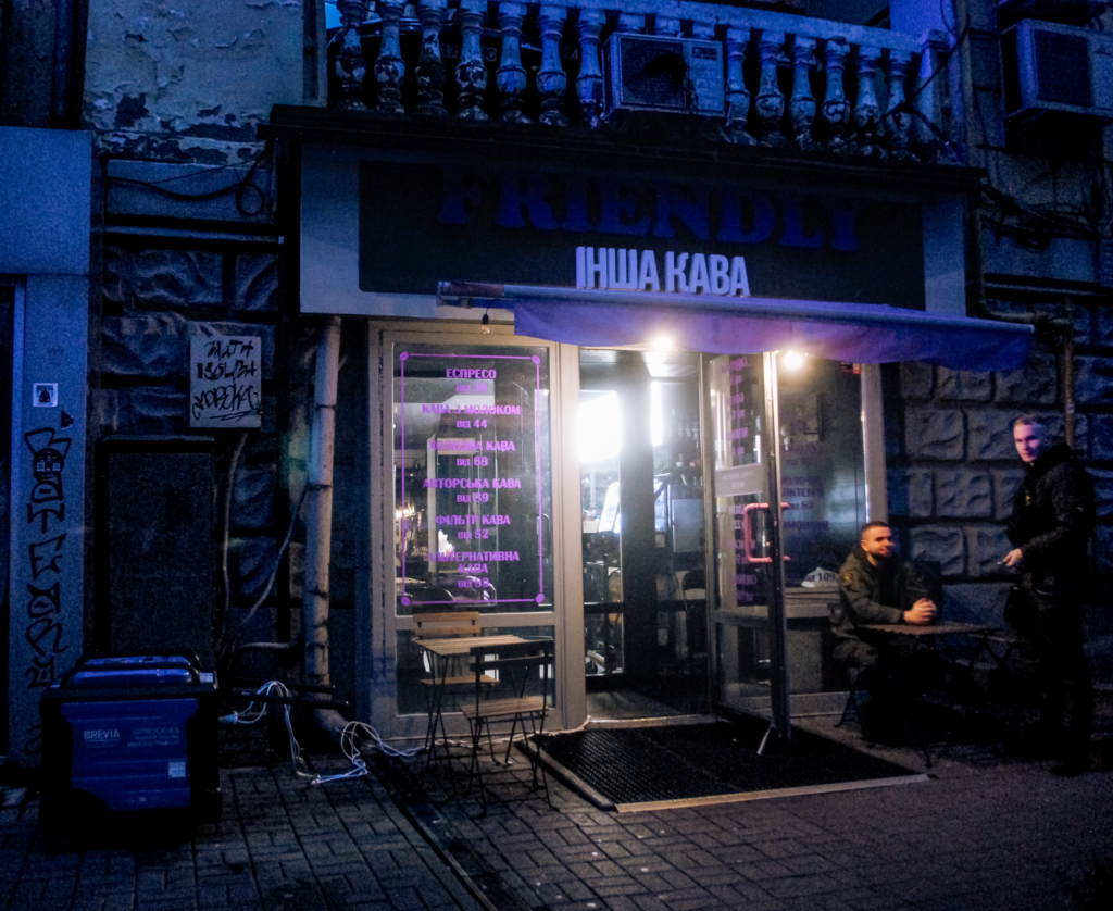 Acoffee shop in Kyiv uses a generator, left, to keep some lights on, and one man sits and another stands at a cafe table to the right of its door.