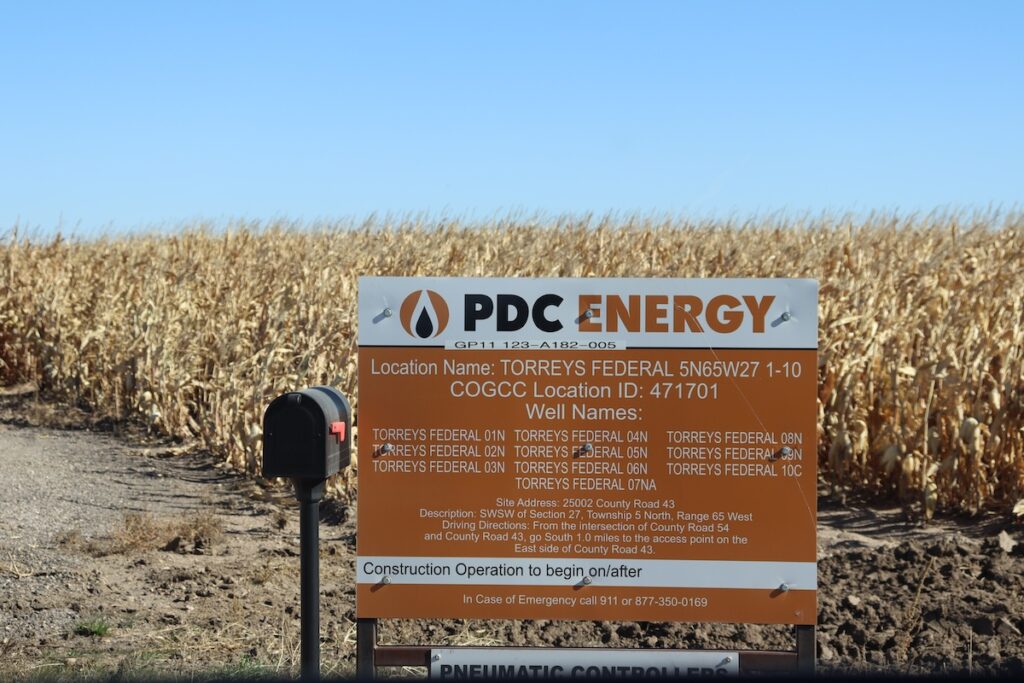 A PDC Energy Energy sign showing nearby oil and gas well sites, in front of a cornfield and mailbox in Weld County, CO.