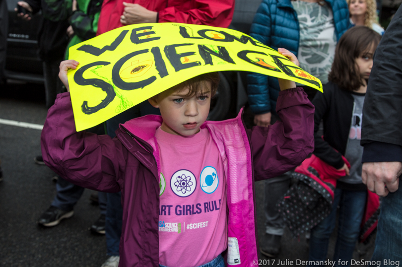 A young girl in a "Smart Girls Rule" t-shirt holds a sign over her head to avoid the rain at the March for Science