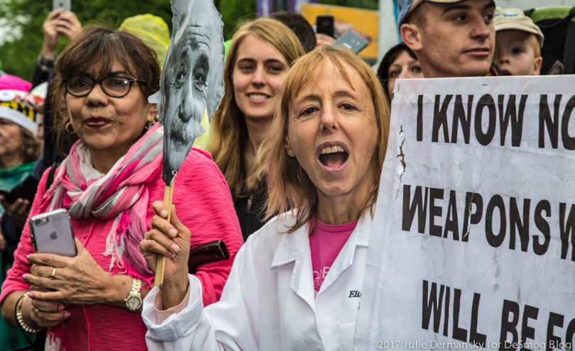 Medea Benjamin, founder of Code Pink, holds an Einstein mask and sign connecting science and war.