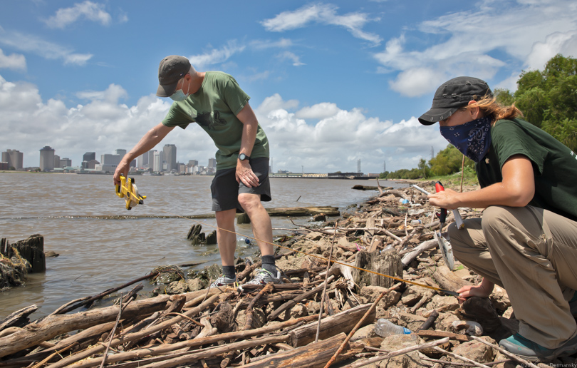 Scientists Mark Benfield and Liz Marchio hold a measuring tape to survey nurdles amid driftwood on a river bank