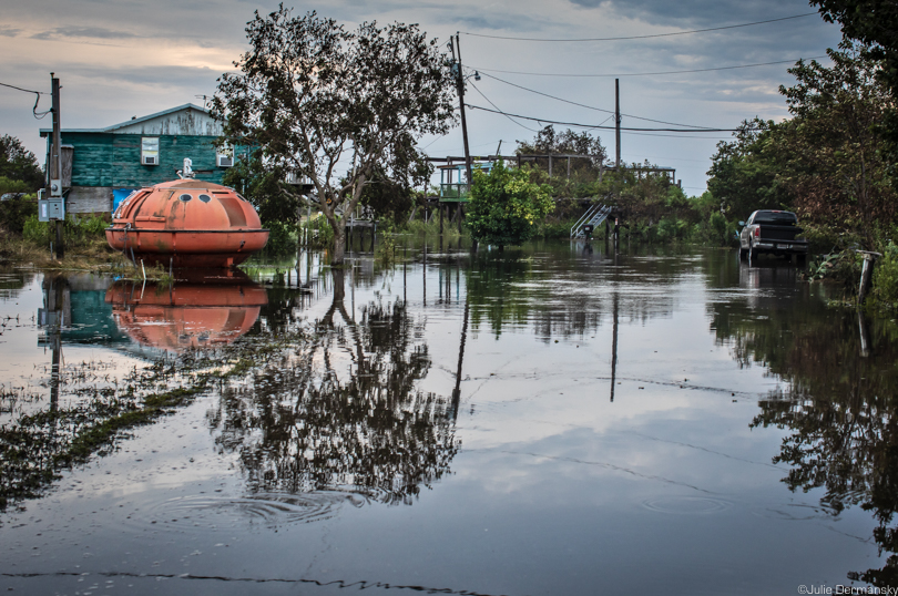 Bright orange oil rig survival pod by a home in standing floodwaters.
