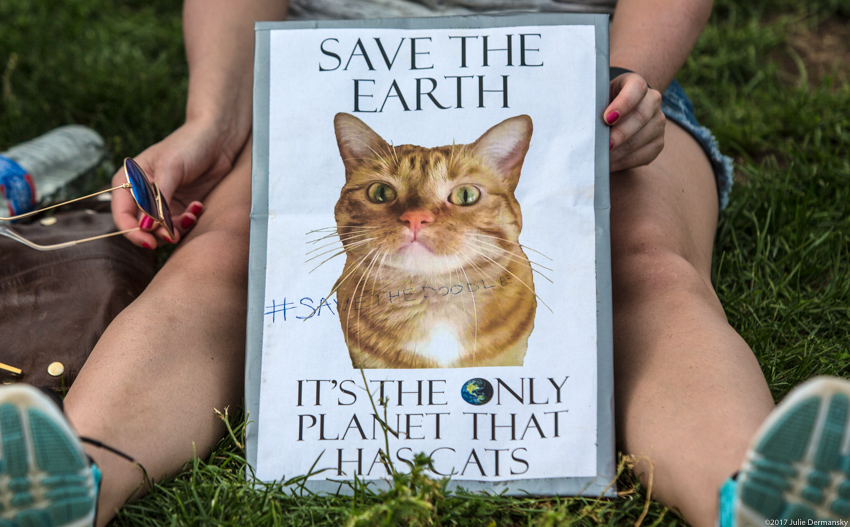 A woman sits with a sign featuring her cat and 'Save the Earth. It's the only planet that has cats.'